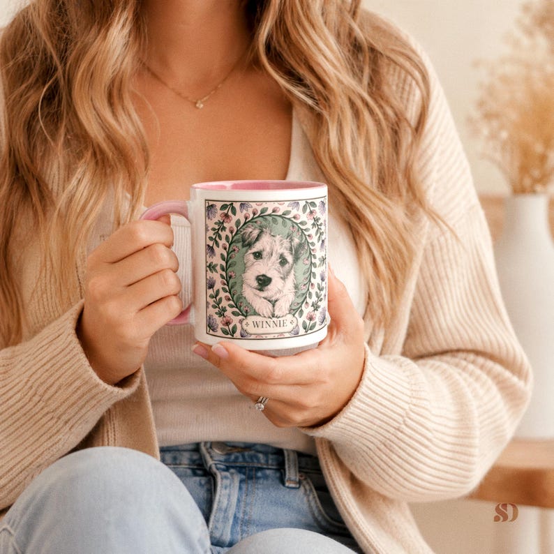 May include: White ceramic mug with a pink handle and rim, featuring a portrait of a dog named "Winnie" surrounded by a floral wreath. The mug is held by a person, showcasing the design and color scheme.