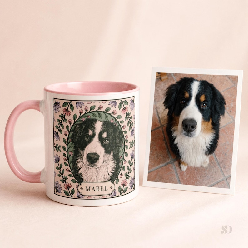 May include: A white ceramic mug with a pink interior and handle, featuring a portrait of a Bernese Mountain Dog named Mabel, surrounded by a floral wreath. A framed photo of the dog sits beside the mug.