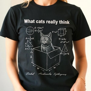May include: A black short-sleeved t-shirt featuring a white graphic of a cat sitting in a box surrounded by mathematical formulas and geometric shapes. The text "What cats really think" appears at the top, with "Euclid", "Archimedes", and "Pythagoras" below.