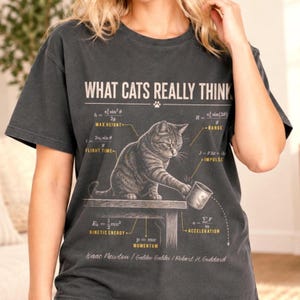 May include: A charcoal grey t-shirt featuring the text WHAT CATS REALLY THINK and a graphic of a tabby cat pushing a mug off a table. The design includes physics equations for max height, range, flight time, impulse, kinetic energy, and acceleration.