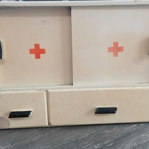 May include: A vintage white medicine cabinet with two doors and two drawers. Each door has a red cross on it. The cabinet has black handles.