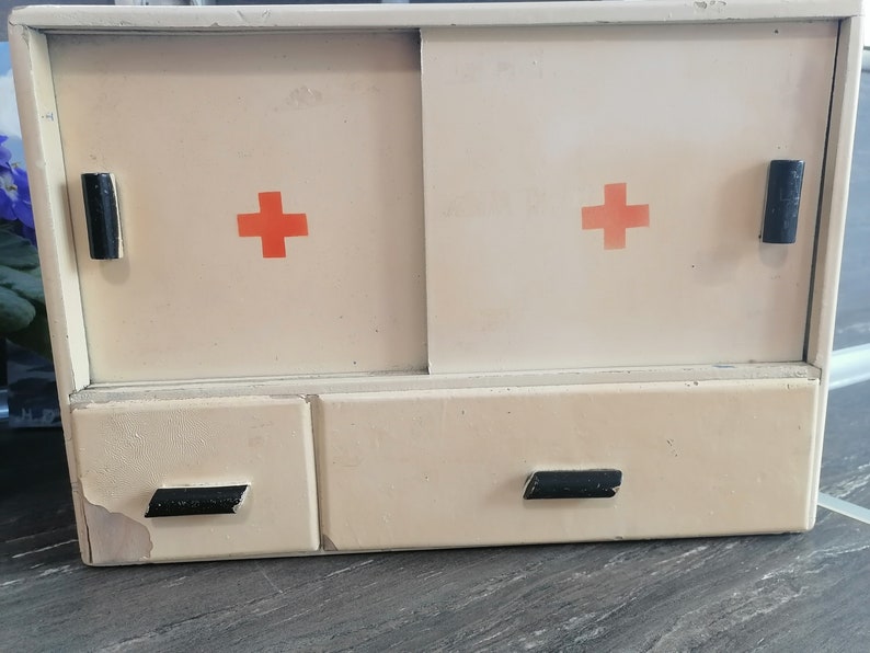 May include: A vintage white medicine cabinet with two doors and two drawers. Each door has a red cross on it. The cabinet has black handles.
