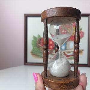 Puede incluir: Un reloj de arena vintage hecho de madera y vidrio. El reloj de arena tiene un marco de madera marr&oacute;n oscuro con una bombilla de vidrio transparente llena de arena blanca. El reloj de arena se sostiene en la mano de una persona.