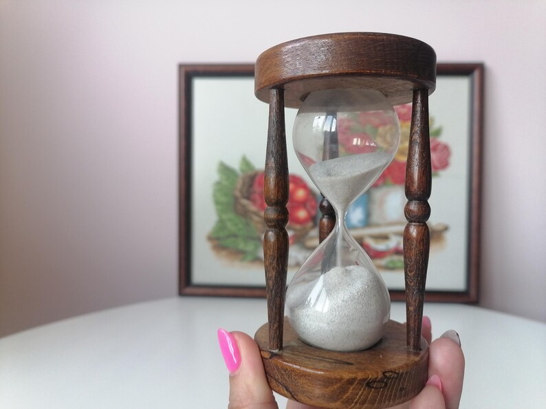 Puede incluir: Un reloj de arena vintage hecho de madera y vidrio. El reloj de arena tiene un marco de madera marr&oacute;n oscuro con una bombilla de vidrio transparente llena de arena blanca. El reloj de arena se sostiene en la mano de una persona.