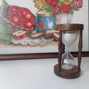 Puede incluir: Un reloj de arena de madera con arena blanca, colocado contra una obra de arte de punto de cruz enmarcada que representa una cesta de manzanas rojas y un arreglo floral. El reloj de arena mide unos 13 cm de alto.
