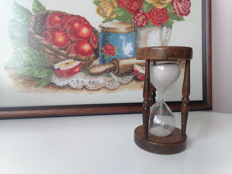 Puede incluir: Un reloj de arena de madera con arena blanca, colocado contra una obra de arte de punto de cruz enmarcada que representa una cesta de manzanas rojas y un arreglo floral. El reloj de arena mide unos 13 cm de alto.