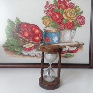Puede incluir: Un reloj de arena de madera con c&aacute;maras de vidrio transparente llenas de arena blanca. El reloj de arena se coloca sobre un fondo de una obra de arte en punto de cruz enmarcada que representa una naturaleza muerta con manzanas y flores. Es un objeto decorativo.
