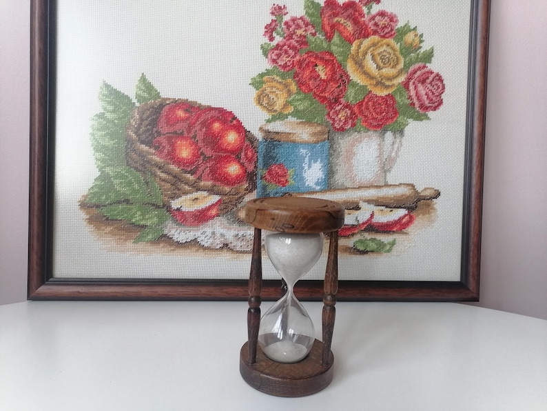 Puede incluir: Un reloj de arena de madera con c&aacute;maras de vidrio transparente llenas de arena blanca. El reloj de arena se coloca sobre un fondo de una obra de arte en punto de cruz enmarcada que representa una naturaleza muerta con manzanas y flores. Es un objeto decorativo.