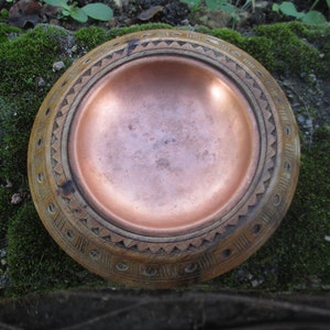 May include: A handcrafted wooden bowl with a copper-colored interior. The rim of the bowl features intricate carved geometric patterns. The bowl is round and appears to be sitting on a bed of green moss.