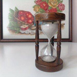 Puede incluir: Un reloj de arena vintage con marco y base de madera. La bombilla de vidrio transparente contiene arena blanca. El reloj de arena est&aacute; sostenido por cuatro pilares de madera oscura. En el fondo, un bordado enmarcado de una cesta de manzanas.