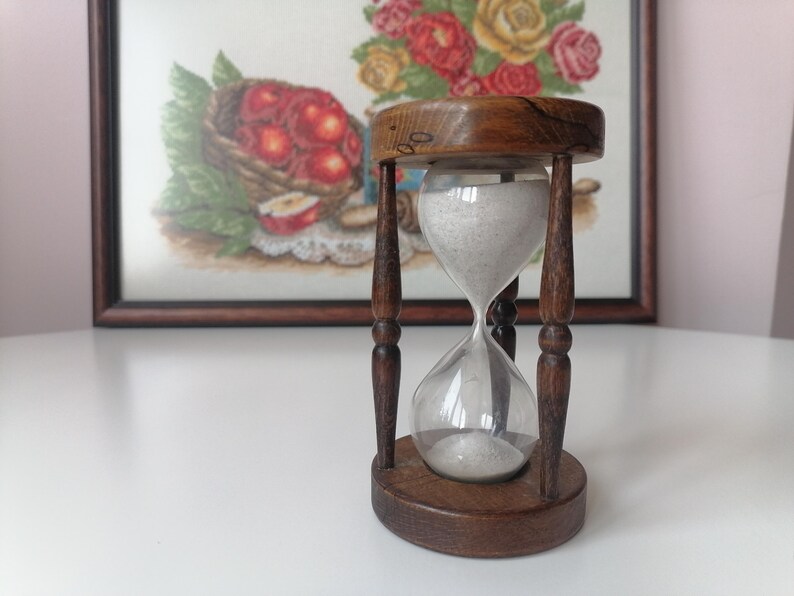 Puede incluir: Un reloj de arena vintage con marco y base de madera. La bombilla de vidrio transparente contiene arena blanca. El reloj de arena est&aacute; sostenido por cuatro pilares de madera oscura. En el fondo, un bordado enmarcado de una cesta de manzanas.