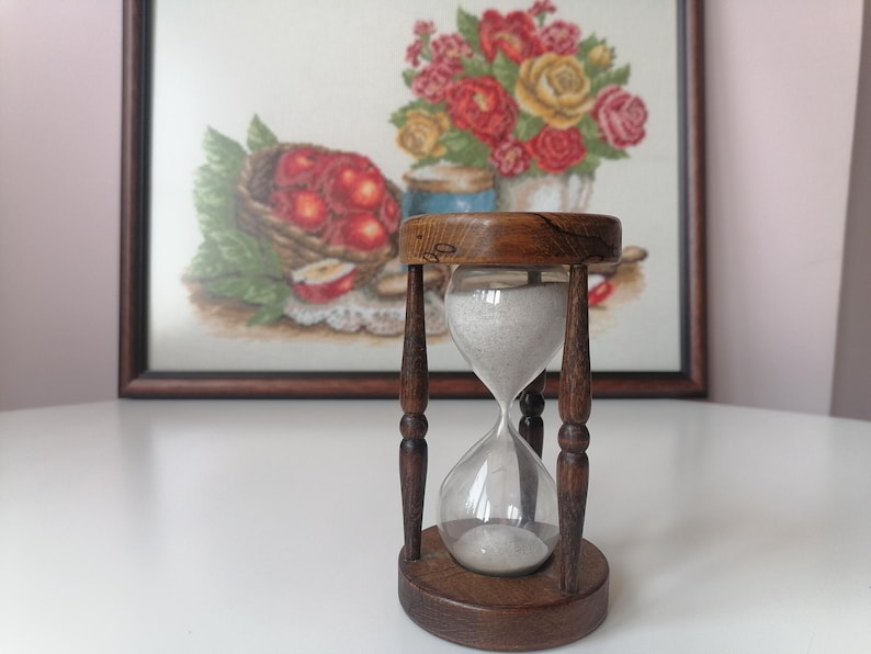 Puede incluir: Un reloj de arena vintage con marco y base de madera. El reloj de arena contiene arena blanca y est&aacute; colocado frente a una pintura enmarcada de flores y frutas. El marco de madera es marr&oacute;n oscuro.
