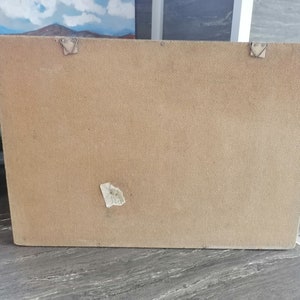 May include: The back of a brown rectangular picture frame with a white label on the bottom right corner. The frame has two metal hooks on the top and two on the bottom.