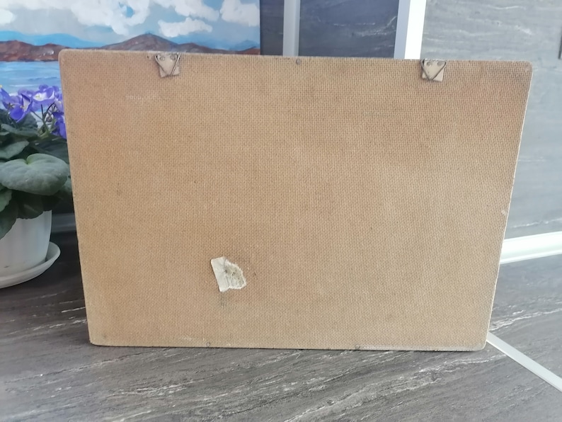 May include: The back of a brown rectangular picture frame with a white label on the bottom right corner. The frame has two metal hooks on the top and two on the bottom.