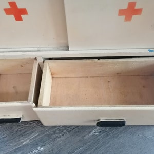 May include: White metal cabinet with two open drawers. The cabinet has a red cross on each side. The drawers are made of wood.