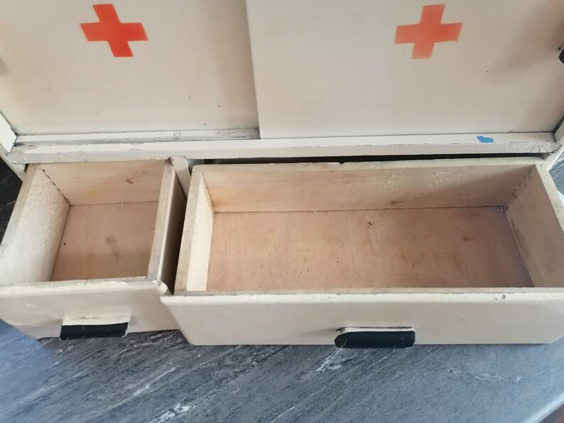 May include: White metal cabinet with two open drawers. The cabinet has a red cross on each side. The drawers are made of wood.