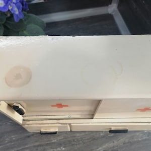 May include: White metal first aid kit with two drawers and red crosses on the front. The kit has two visible water stains on the top.