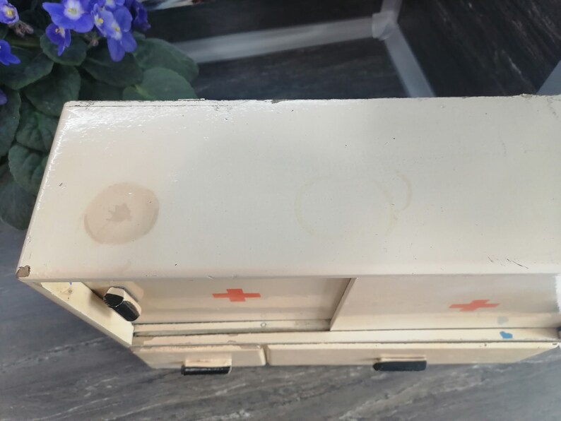 May include: White metal first aid kit with two drawers and red crosses on the front. The kit has two visible water stains on the top.