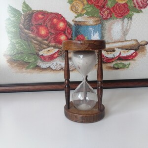 Puede incluir: Un reloj de arena de madera vintage con un centro de vidrio transparente, lleno de arena blanca. El reloj de arena se encuentra frente a una obra de arte en punto de cruz enmarcada que representa una naturaleza muerta de manzanas y flores. El marco y la base de madera tienen un acabado marr&oacute;n oscuro.