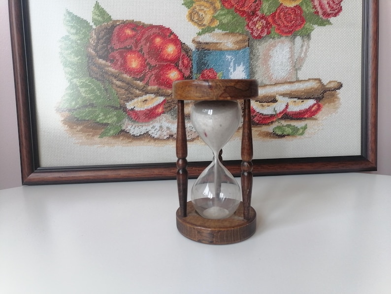 Puede incluir: Un reloj de arena de madera vintage con un centro de vidrio transparente, lleno de arena blanca. El reloj de arena se encuentra frente a una obra de arte en punto de cruz enmarcada que representa una naturaleza muerta de manzanas y flores. El marco y la base de madera tienen un acabado marr&oacute;n oscuro.