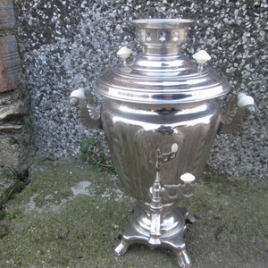 May include: A silver samovar with white porcelain accents. The samovar has a round body with a spout and a handle on each side. It sits on a four-legged base.