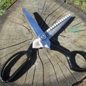 May include: A pair of silver-colored fabric shears with black handles. The shears are open and lying on a wooden surface.