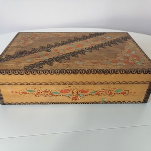 Puede incluir: Una caja de madera con un diseño decorativo. La caja está pintada con un patrón floral en tonos naranja, azul y verde. La caja tiene un borde marrón oscuro alrededor de los bordes.