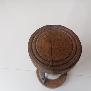 Puede incluir: Un reloj de arena vintage de madera marr&oacute;n oscuro. La parte superior e inferior son circulares, con un dise&ntilde;o tallado alrededor de los bordes. El reloj de arena tiene un tallo estrecho y curvo y un centro de vidrio transparente.