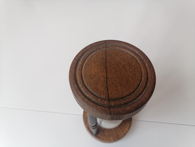 Puede incluir: Un reloj de arena vintage de madera marr&oacute;n oscuro. La parte superior e inferior son circulares, con un dise&ntilde;o tallado alrededor de los bordes. El reloj de arena tiene un tallo estrecho y curvo y un centro de vidrio transparente.