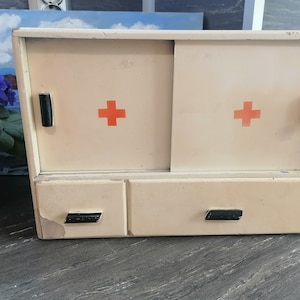 May include: A vintage white medicine cabinet with two doors and two drawers. The doors have red crosses on them and black handles. The cabinet has a worn, distressed look.