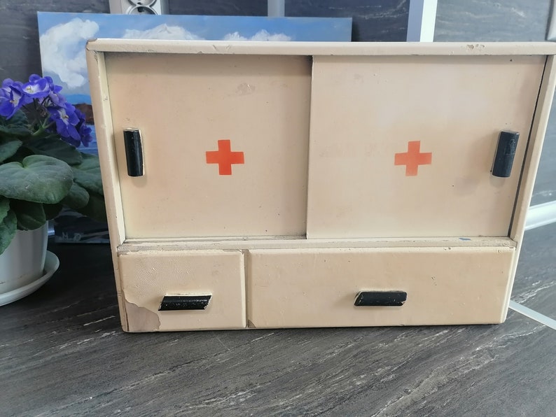 May include: A vintage white medicine cabinet with two doors and two drawers. The doors have red crosses on them and black handles. The cabinet has a worn, distressed look.