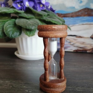 Könnte beinhalten: Eine Sanduhr aus Holz mit weißem Sand, die auf einer dunkelgrauen Oberfläche steht. Die Sanduhr ist scharf, während der Hintergrund unscharf ist.