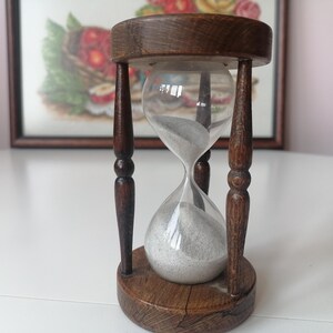 Puede incluir: Un reloj de arena vintage con marco y base de madera. La bombilla de vidrio transparente contiene arena gris. El marco de madera es de color marr&oacute;n oscuro y tiene un dise&ntilde;o decorativo. El reloj de arena est&aacute; sobre una superficie blanca.