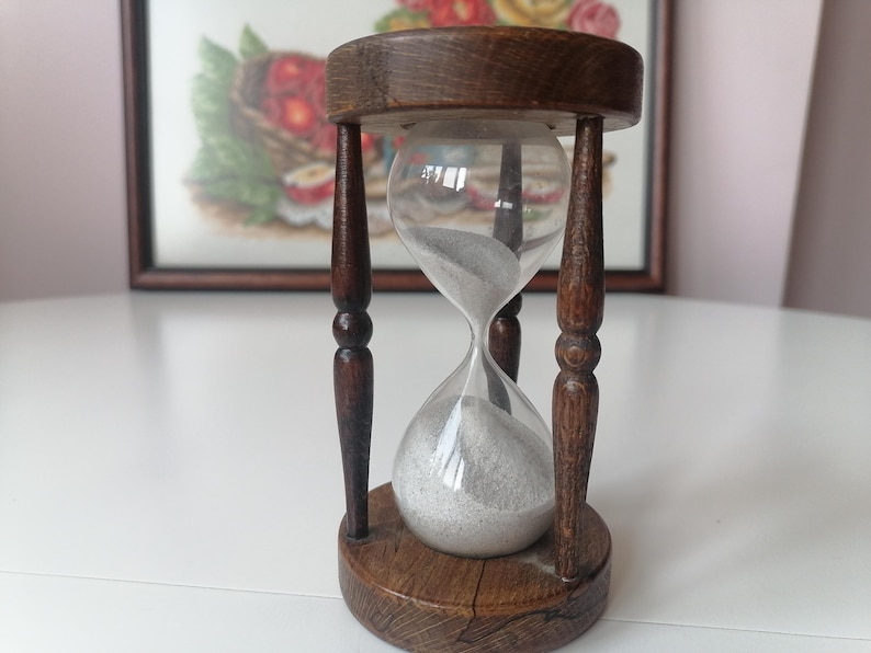 Puede incluir: Un reloj de arena vintage con marco y base de madera. La bombilla de vidrio transparente contiene arena gris. El marco de madera es de color marr&oacute;n oscuro y tiene un dise&ntilde;o decorativo. El reloj de arena est&aacute; sobre una superficie blanca.