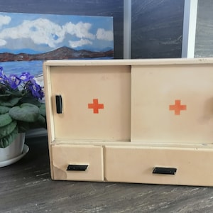 May include: A white vintage medicine cabinet with two doors and a drawer. The doors have red crosses on them and black handles. The drawer has two black handles.