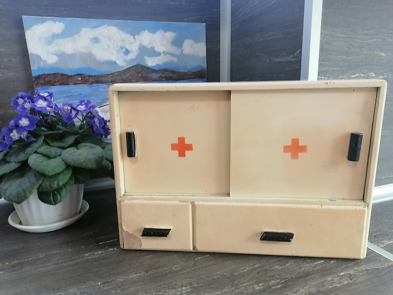 May include: A white vintage medicine cabinet with two doors and a drawer. The doors have red crosses on them and black handles. The drawer has two black handles.