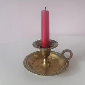 Puede incluir: Un candelero de latón vintage con una vela roja. El candelero tiene una base redonda con un borde decorativo y un asa pequeña. La vela mide aproximadamente 15 cm de altura.