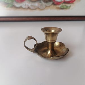 May include: A vintage brass candle holder with a handle. The candle holder has a small, round base and a flared cup to hold a candle. The handle is a small loop. The candle holder is a warm, golden colour.