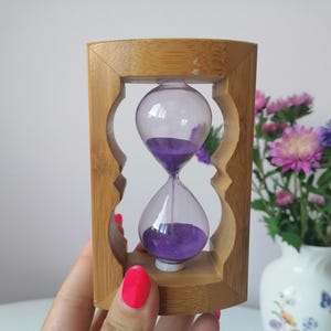 Könnte beinhalten: Eine Sanduhr aus Holz mit einem violetten Sandtimer. Die Sanduhr befindet sich in einem Holzrahmen mit einem dekorativen Design.