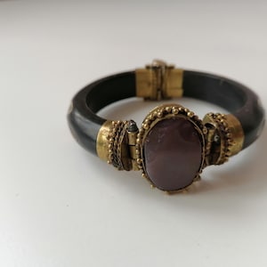 Op de afbeelding: Een zwarte bangle armband met een gouden sluiting en een grote, ovale, paarse steen in een gouden frame.