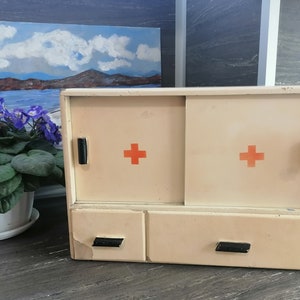 May include: A white medicine cabinet with two doors and a drawer. The doors have red crosses on them. The cabinet is made of wood and has a vintage look.