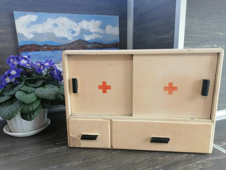 May include: A white medicine cabinet with two doors and a drawer. The doors have red crosses on them. The cabinet is made of wood and has a vintage look.