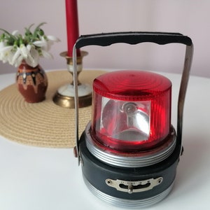 May include: A vintage red and black lantern with a handle and a clear glass lens. The lantern has a silver band around the middle and a switch on the side.