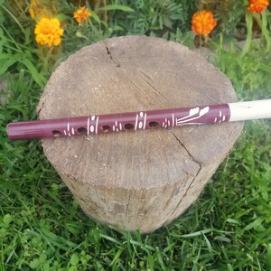 May include: A cylindrical wooden flute with a deep maroon finish, featuring white painted floral and linear patterns along its length. The instrument has a series of circular finger holes and a light-colored, unpainted wooden mouthpiece at one end.