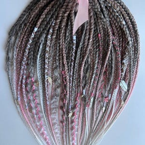 Könnte beinhalten: Ein Set synthetischer Haarverlängerungen in Braun-, Rosa- und Weißtönen. Die Verlängerungen sind geflochten und haben kleine Charms angebracht. Die Verlängerungen sind mit einem rosa Band zusammengebunden.
