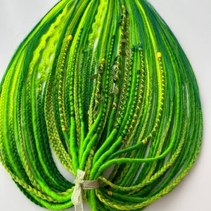 Puede incluir: Un paquete de rastas sintéticas verdes y amarillas con detalles verdes y amarillos. Las rastas están atadas con una cinta plateada.