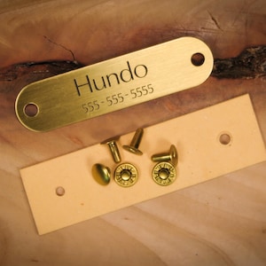 Op de afbeelding: Een messing hondenpenning met de naam "Hundo" en een telefoonnummer gegraveerd, samen met een lichtbruine leren strip en verschillende messing klinknagels. De items zijn gerangschikt op een houten oppervlak.