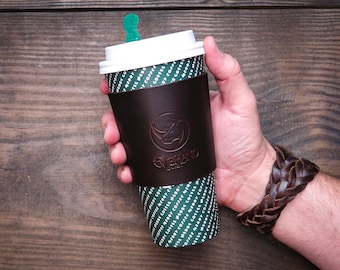 May include: A brown leather coffee cup holder with a rhino logo and the text "Overland" on it. The holder is wrapped around a disposable coffee cup with a white lid and a green straw.