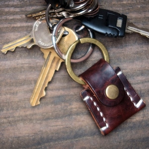 May include: A brown leather key chain with a stitched border and a brass button. The keychain is attached to a set of keys with a brass ring.
