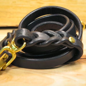 Op de afbeelding: Een zwarte leren riem met een gevlochten gedeelte en een gouden metalen gesp.